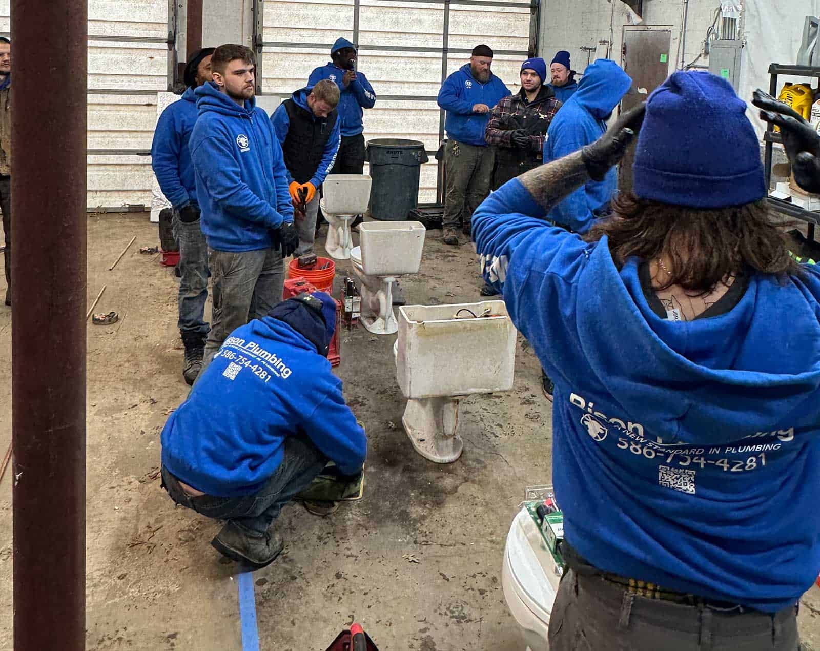 A group of workers in blue hoodies with a plumbing company logo gather inside a workshop, some kneeling or standing near toilets on the floor, preparing tools and equipment. The space appears busy and industrial.
