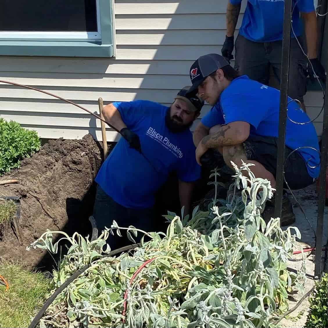 Two plumbers in blue shirts work outside a house; one kneels in a hole near the foundation, while the other crouches beside him. Plants and soil are in the foreground, and a window is visible on the house.