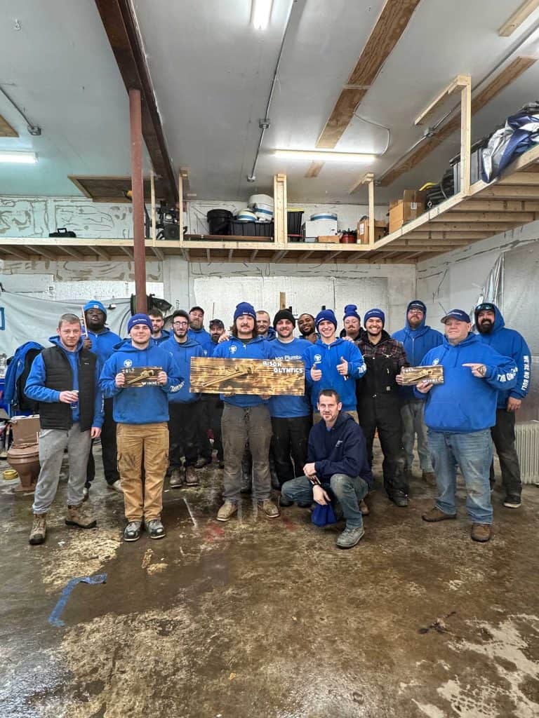A group of people in blue hoodies pose together in a workshop, smiling and holding wooden signs. Some are standing, some kneeling, and tools and shelves are visible in the background. The floor appears wet.