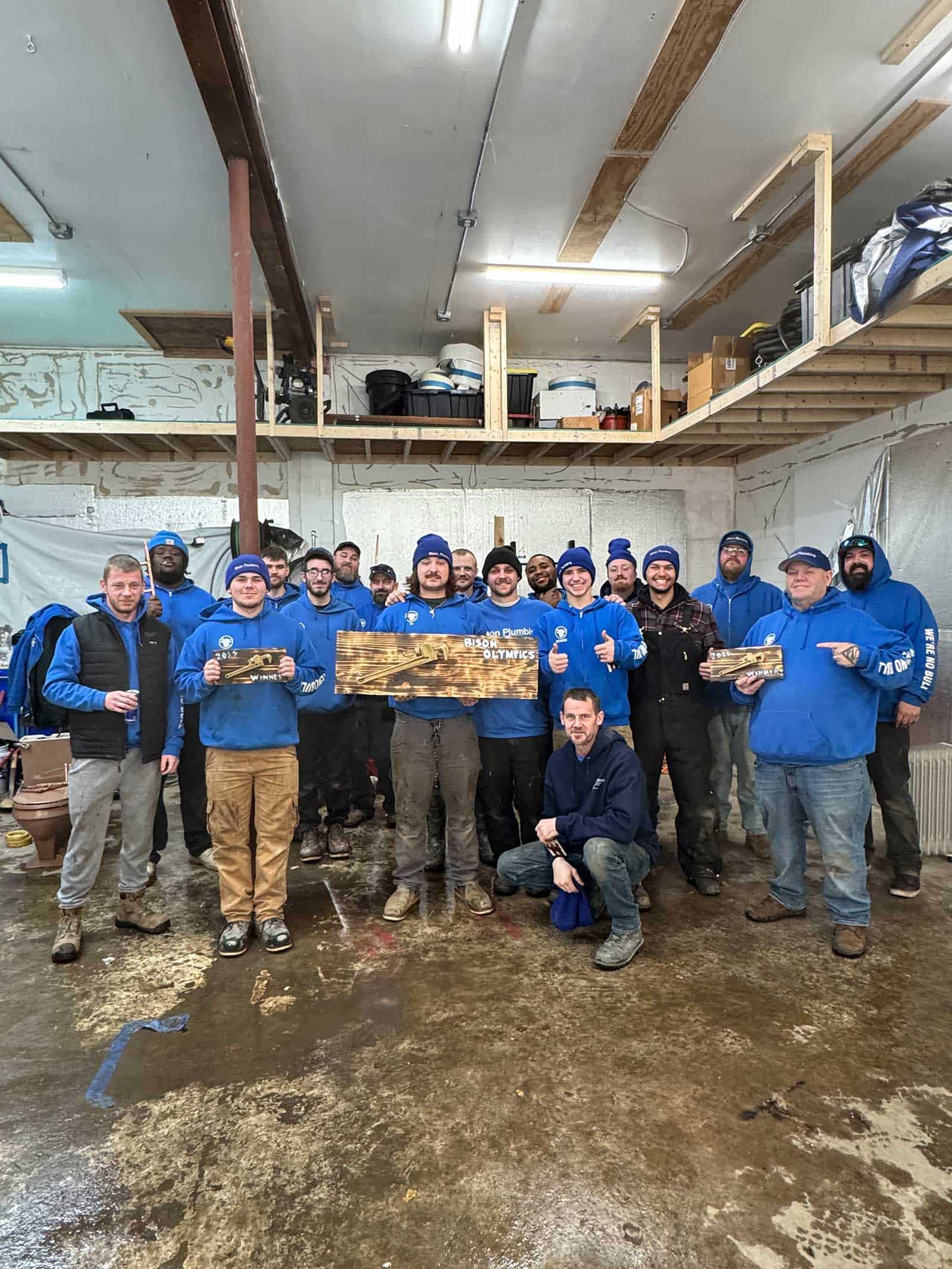 A group of people in blue hoodies pose together in a workshop, smiling and holding wooden signs. Some are standing, some kneeling, and tools and shelves are visible in the background. The floor appears wet.