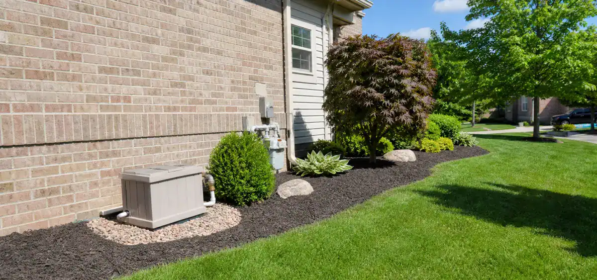 A landscaped yard with a brick house, green grass, shrubs, rocks, mulch, and small generator or utility box near the wall. Bright, sunny day with blue sky and some trees in the background.