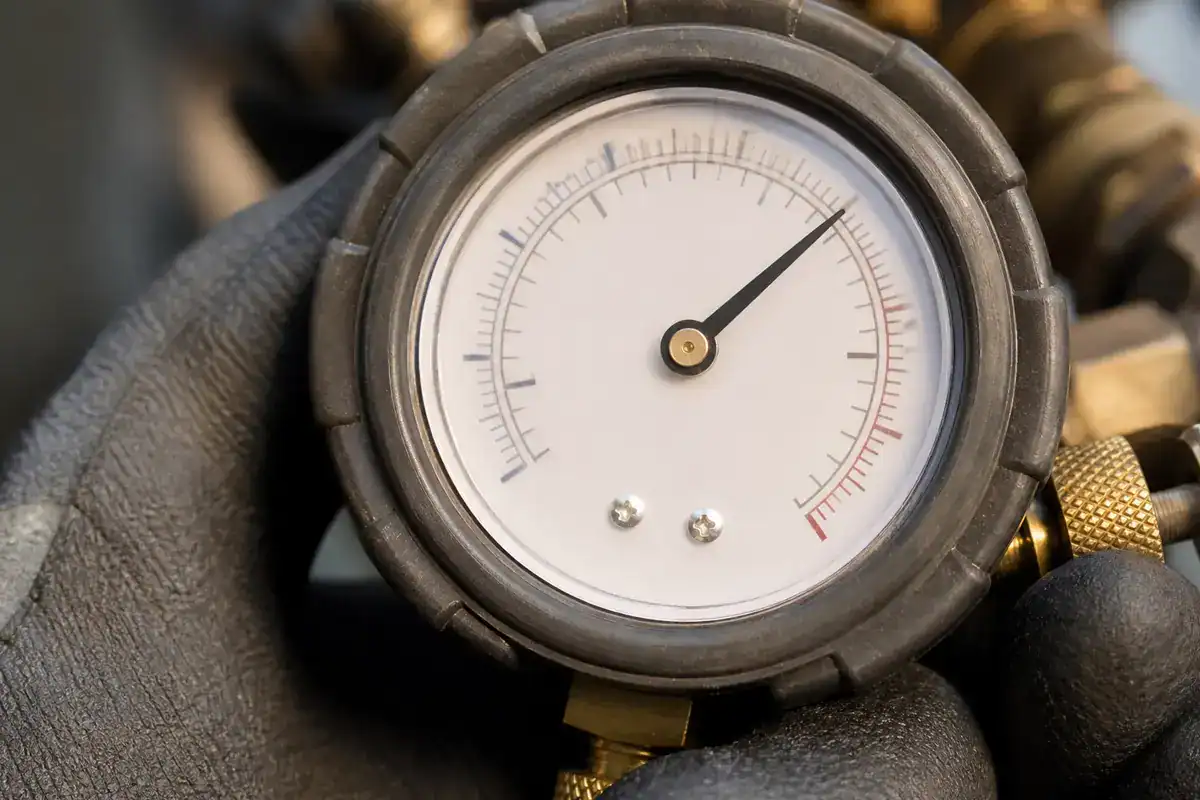 A close-up of a gloved hand holding a round pressure gauge, with the needle pointing to a mid-range value on the dial, showing both black and red scale markings.