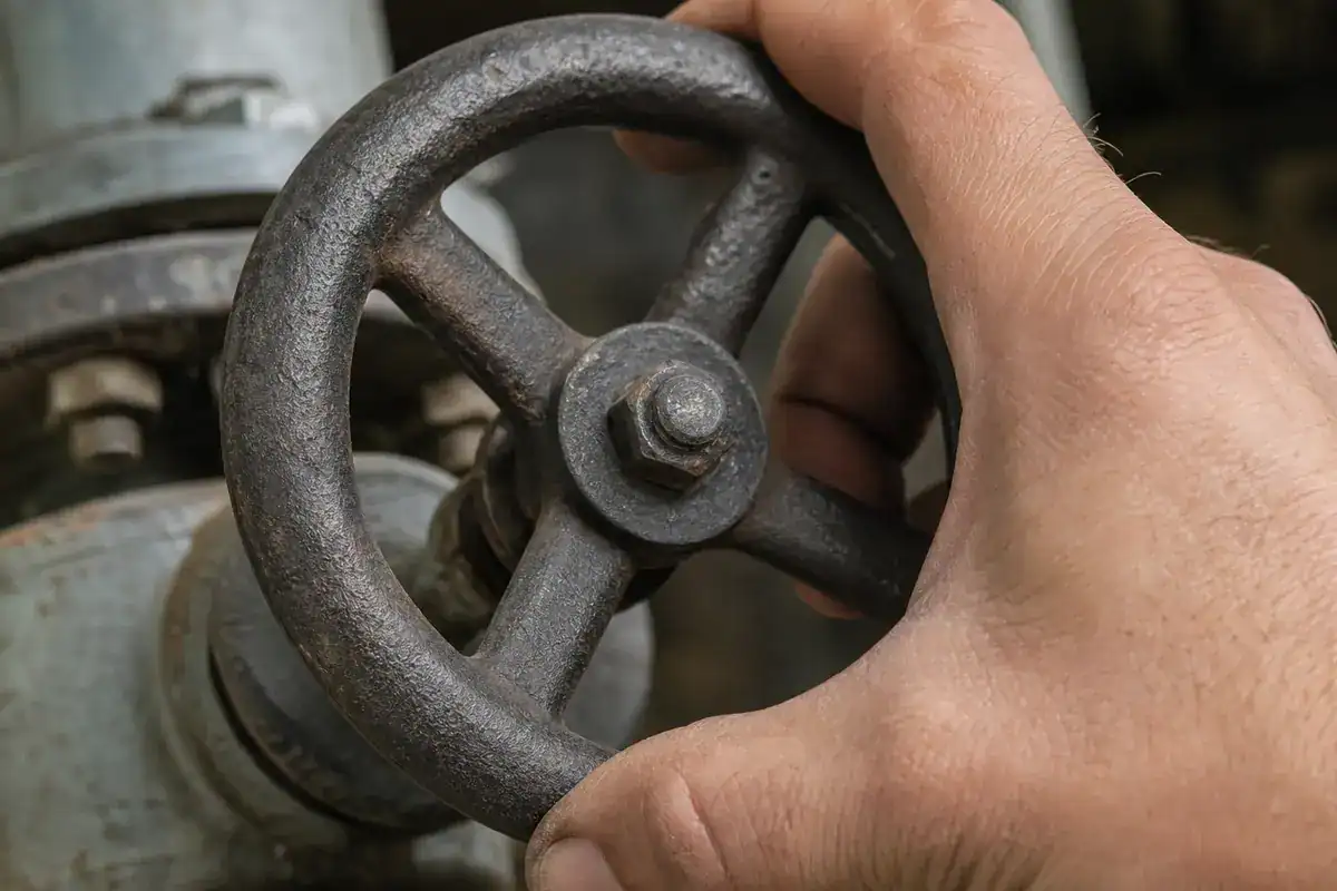 A hand turns a rusty metal valve wheel, which is attached to a pipe.