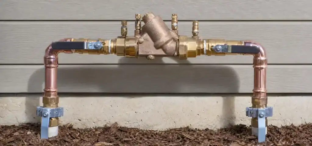 A brass backflow preventer is connected to copper pipes, mounted against a houses exterior wall above mulch and soil.