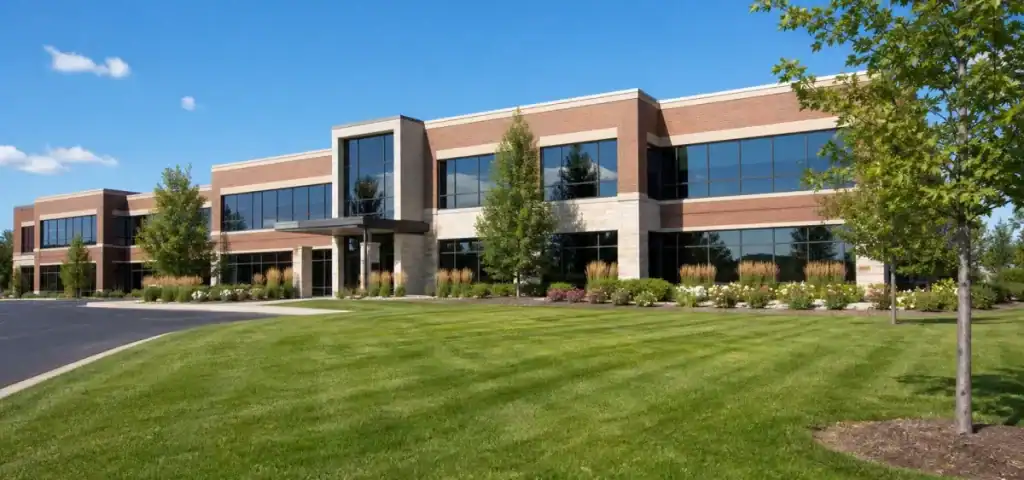 A modern two-story office building with large glass windows, surrounded by green grass, landscaping, and trees under a clear blue sky.
