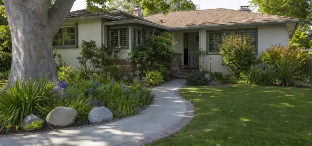 A single-story house with beige walls, a brown shingle roof, large front windows, and a curved walkway surrounded by green grass, bushes, flowers, and a large tree in the front yard.
