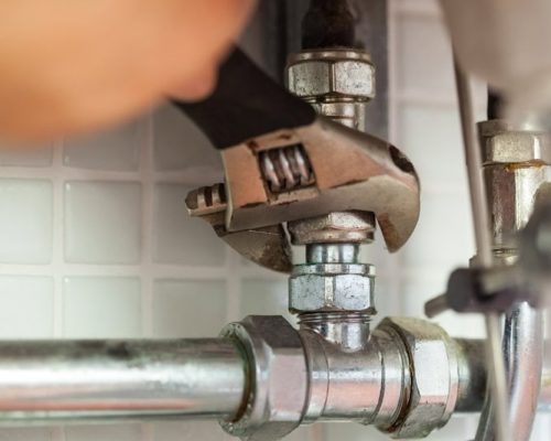 A close-up of a hand using a wrench to tighten a metal pipe fitting under a sink, with tiled wall in the background—perfect for anyone searching "plumber near me" or considering Bison Plumbing Warren Michigan for expert service.